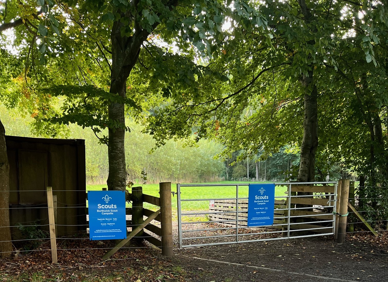 Entrance to Borthwick Scout Campsite: a metal field gate and wooden fence under tall trees, with blue Scouts Borthwick Scout Campsite signs, and a grassy field beyond
